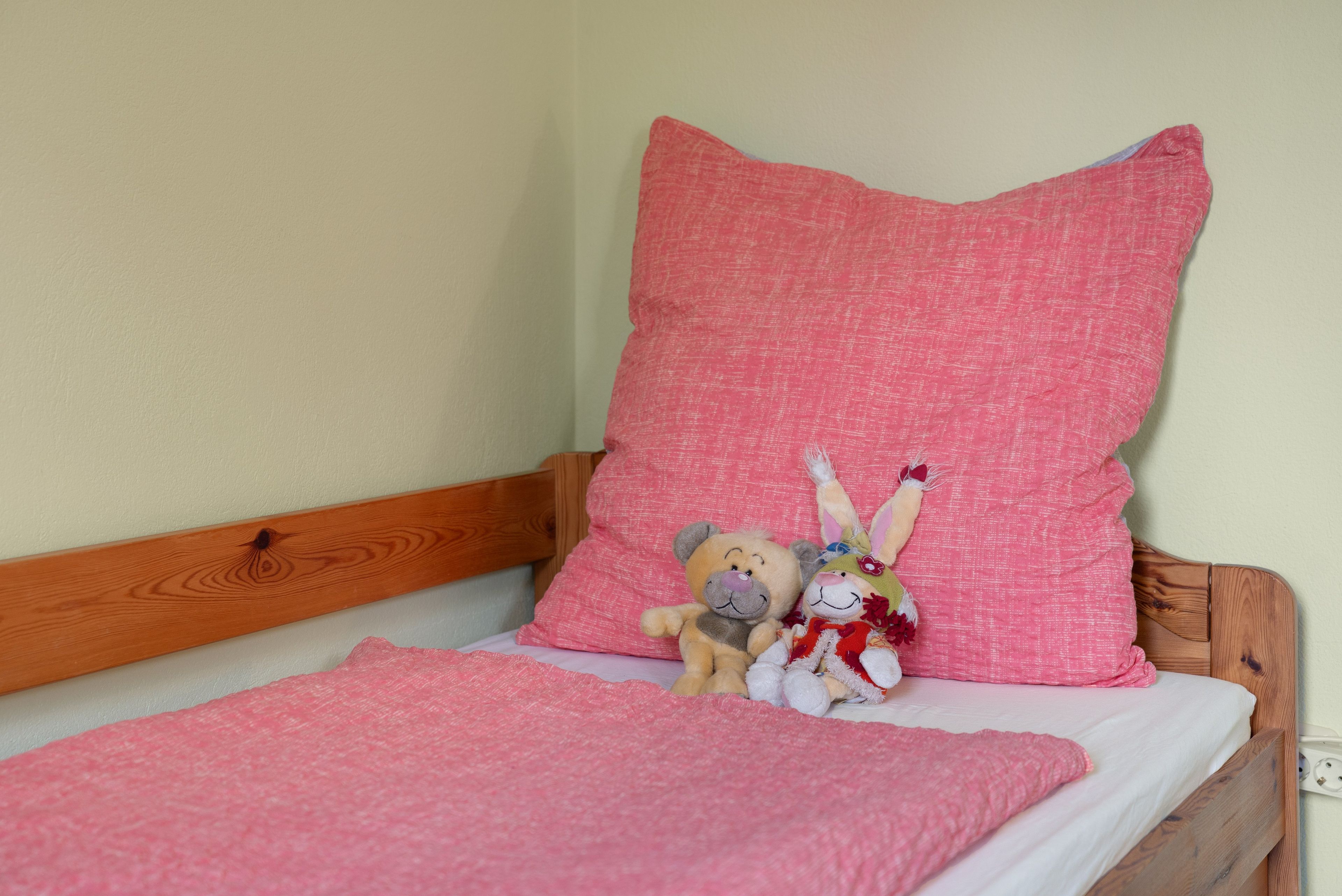 Ein Bett mit einer rosa Decke und einem Kopfkissen, einem hölzernen Kopfteil und zwei Stofftieren auf dem Bett.
