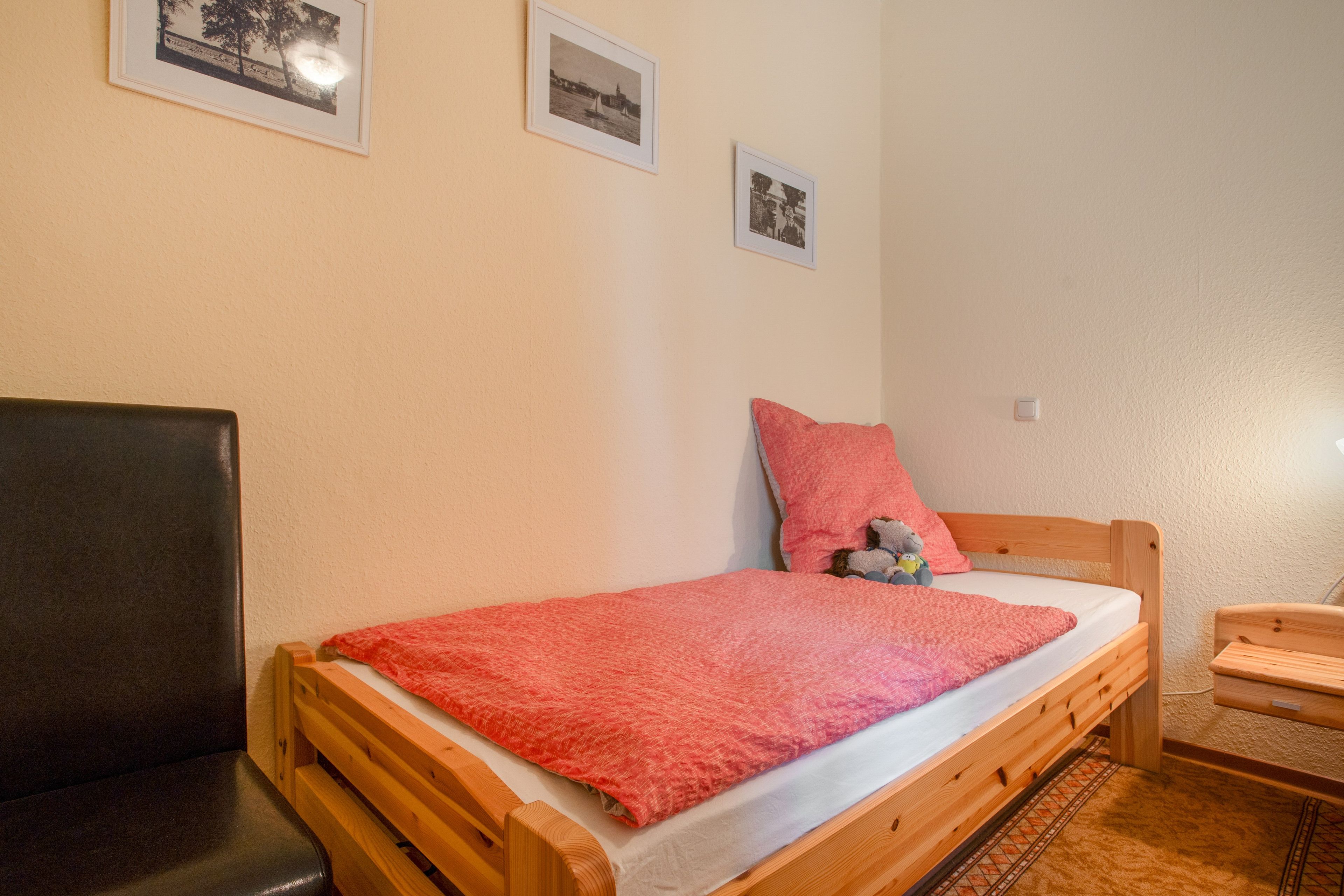 A single bed with a red blanket, a wooden headboard, and a small bedside table with a lamp. There are three framed pictures on the wall.
