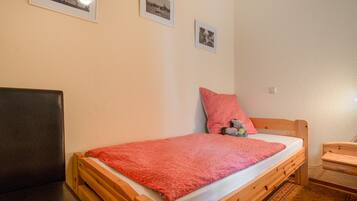 A single bed with a red blanket, a wooden headboard, and a small bedside table with a lamp. There are three framed pictures on the wall.
