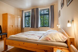 Une chambre à coucher avec un lit en bois, deux fenêtres avec des rideaux, un radiateur, une armoire en bois et une chaise.