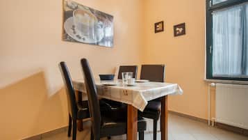 A dining area with a table set for two, chairs, a window with curtains, and a wall with framed pictures.