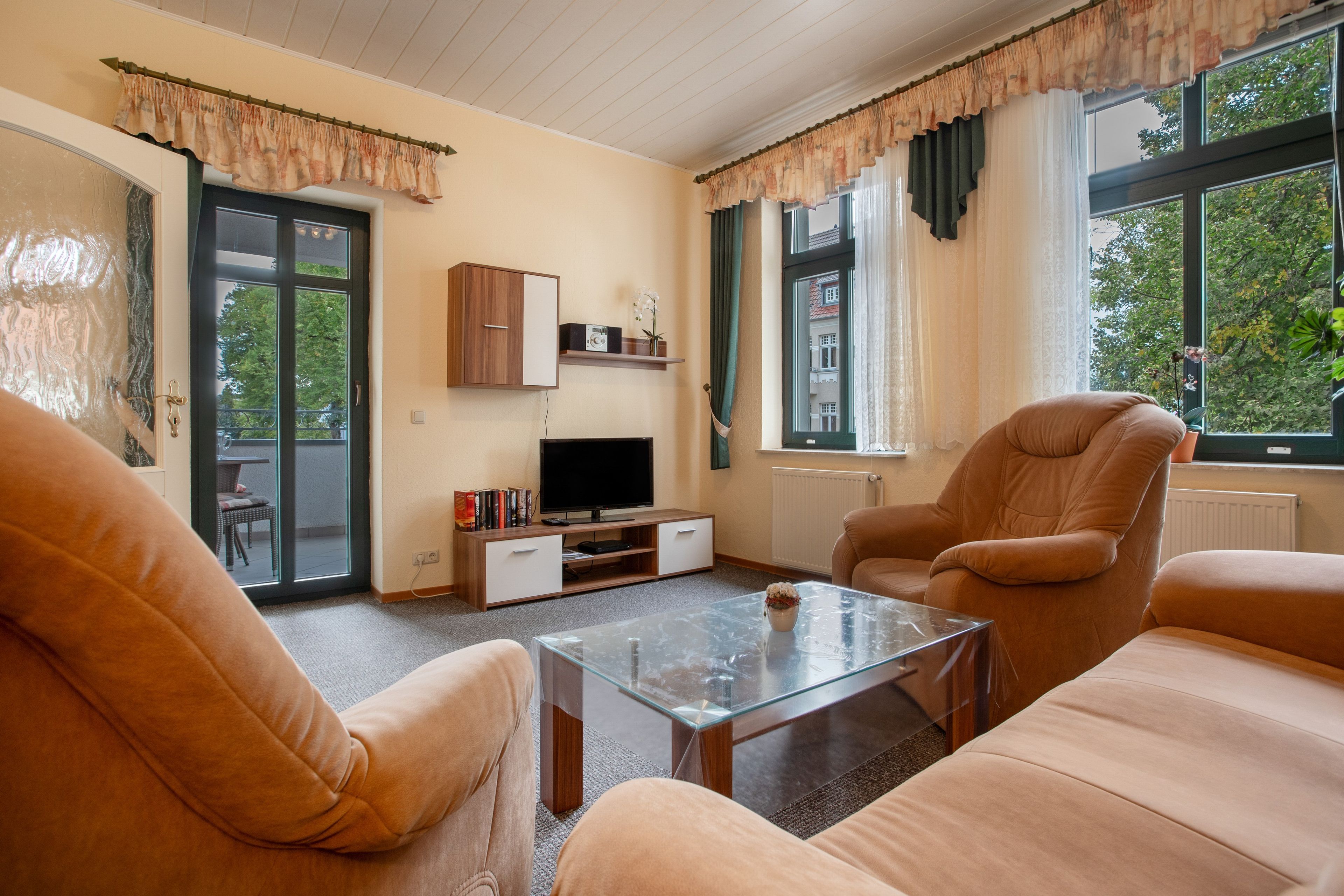 Ein Wohnzimmer mit Sofa, Sessel, Glastisch, TV-Möbel und Bücherregal.