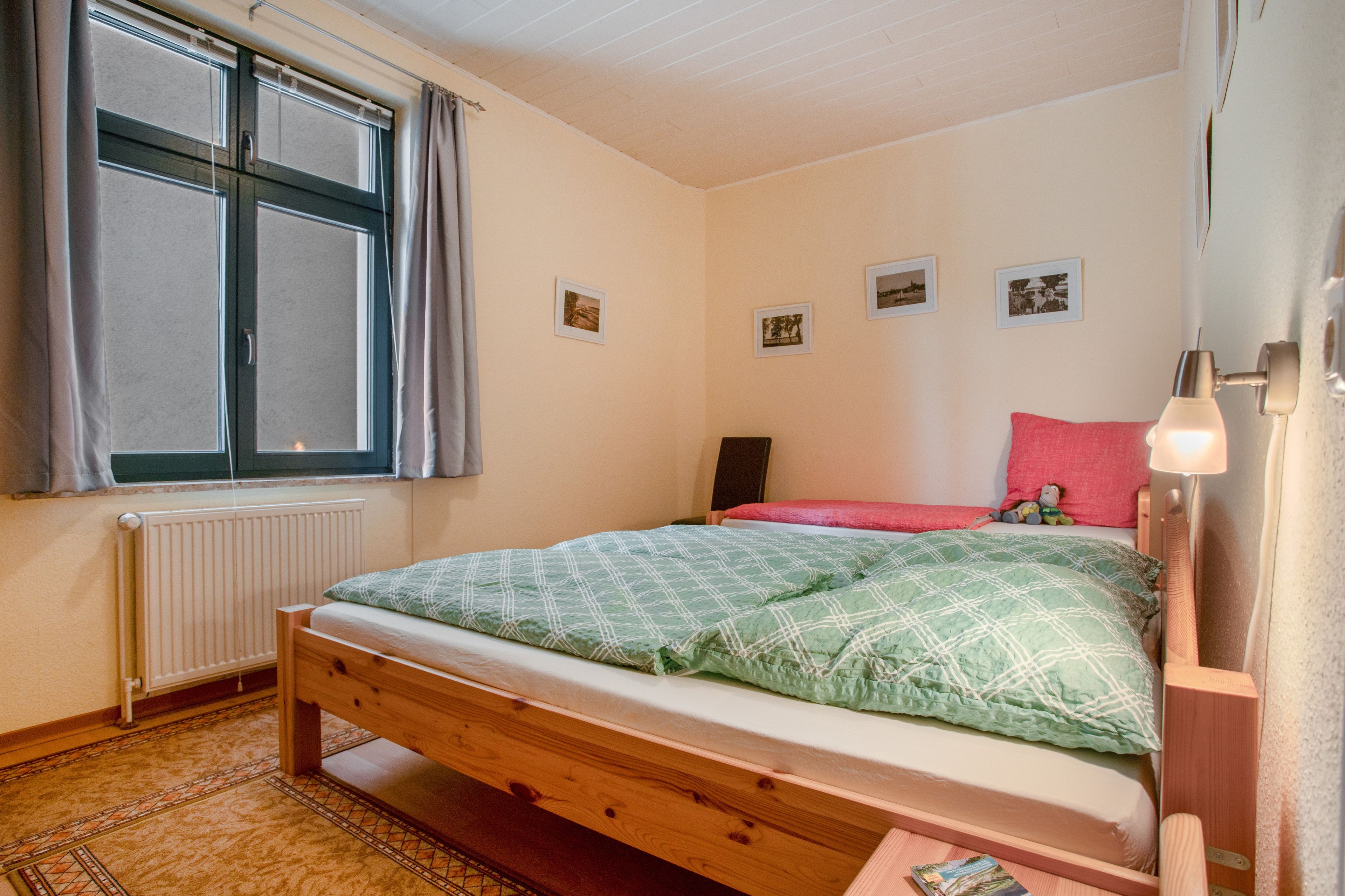 A bedroom with a single bed, a window with curtains, a radiator, and a wall with three framed pictures.