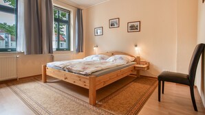 Ein Schlafzimmer mit einem hölzernen Bett, einem Sessel, einem Nachttisch, einem Fenster mit Vorhängen und zwei Wandlampen.