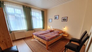 A bedroom with a wooden bed, two bedside tables, a chair, a window with curtains, and three framed pictures on the wall.