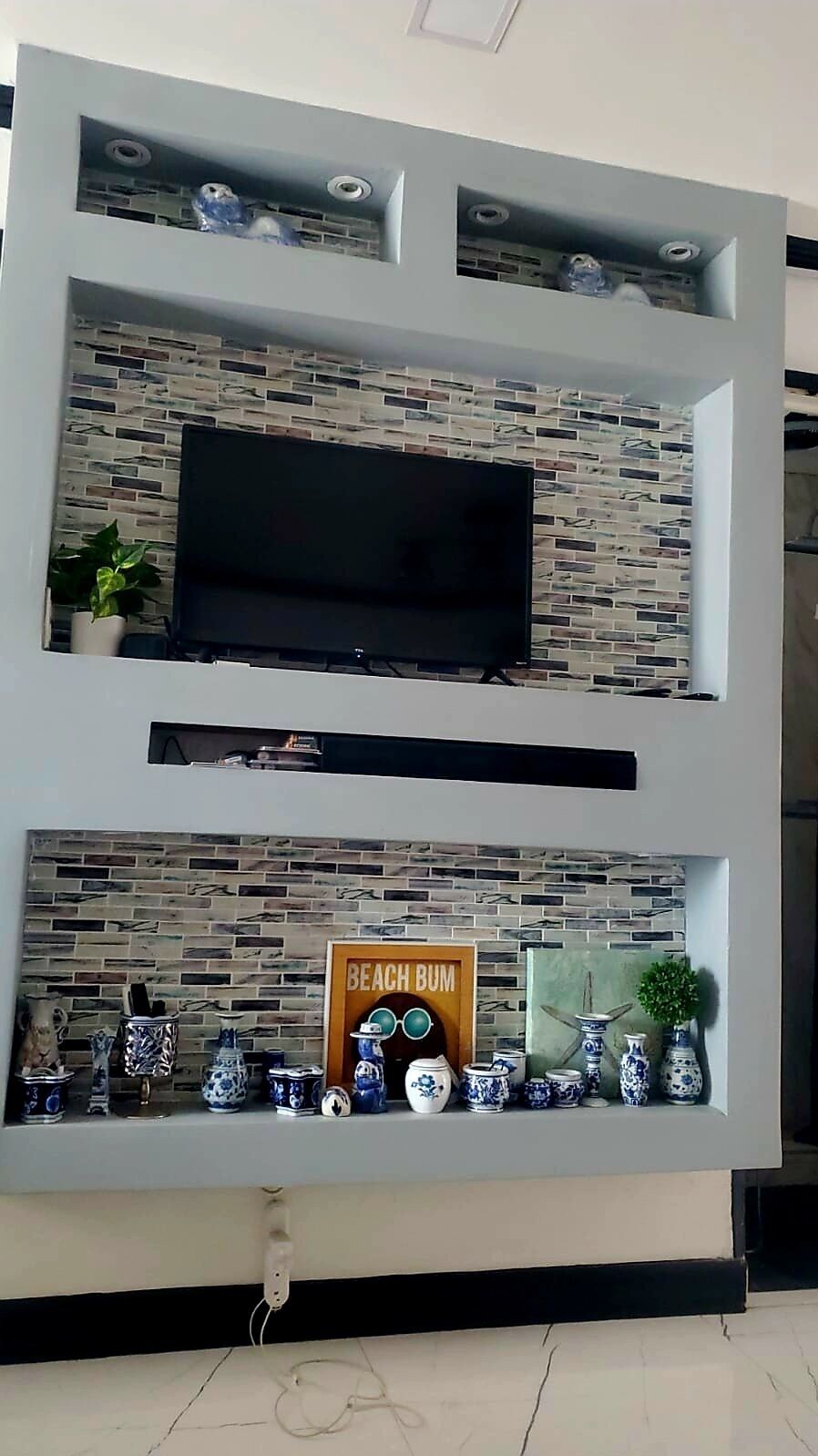 A modern living room with a TV mounted on a brick-patterned wall, a shelf with decorative items below, and a 'Beach Bum' sign.