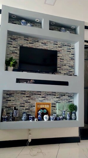 Ein modernes Wohnzimmer mit an einer Ziegelwand montiertem Fernseher, einem darunterliegenden Regal mit Dekoration und einem Schild mit der Aufschrift 'Beach Bum'.
