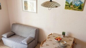 A room with a grey sofa, a dining table with a heart-patterned tablecloth, a bowl of fruit, and a small plant. - Mountain views and comfort near Barcelonnette (Enchastrayes)