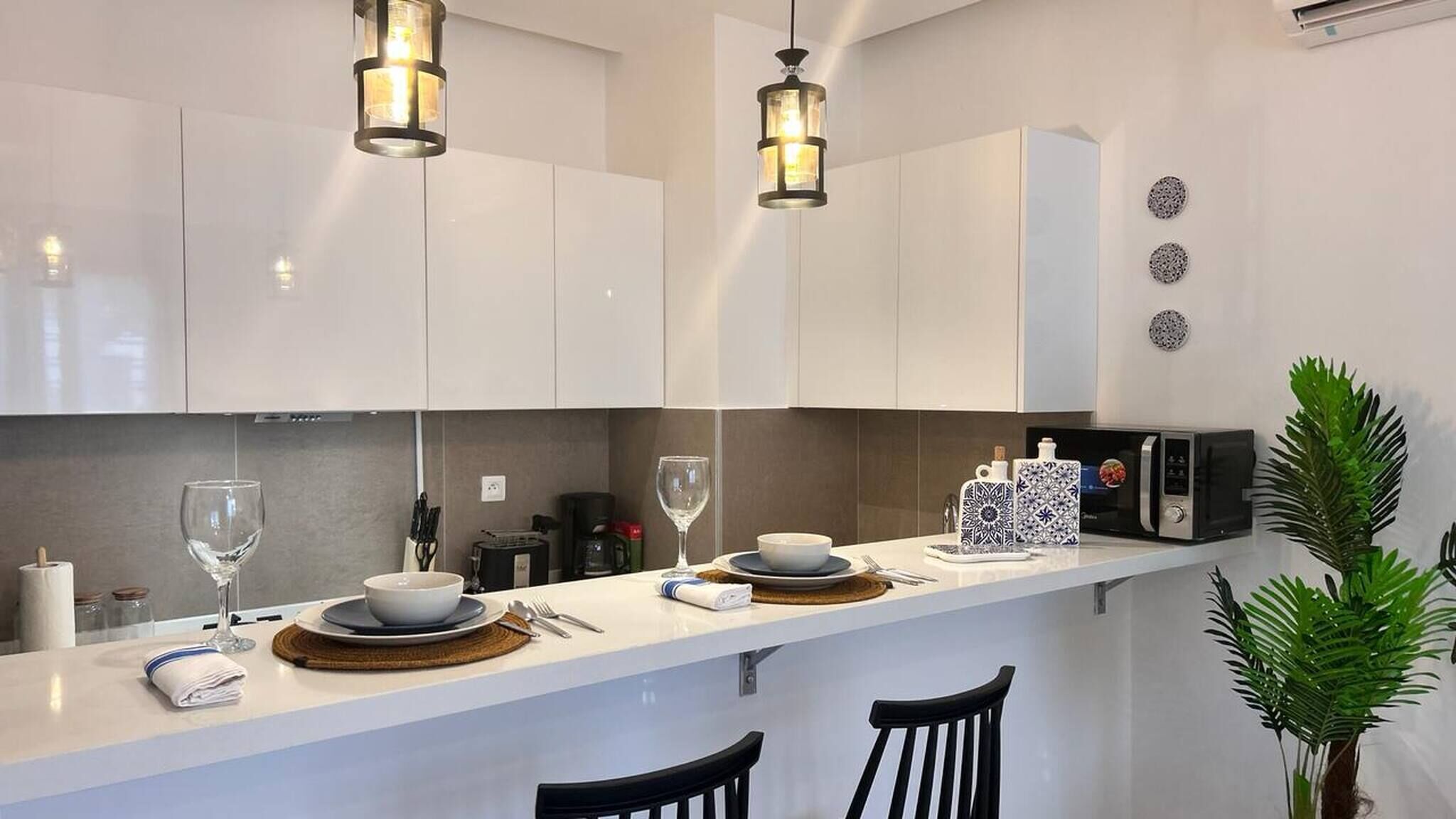 Une cuisine moderne avec des meubles blancs, un plan de travail, deux suspensions lumineuses, un four à micro-ondes et une plante en pot.