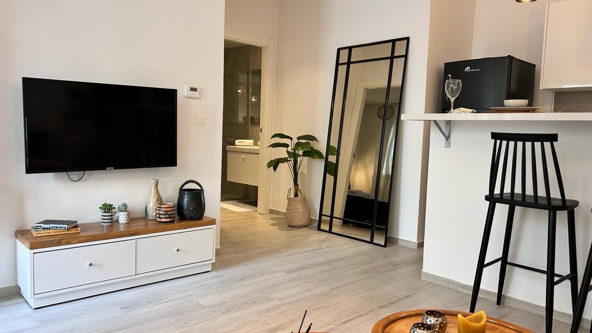 Un salon moderne avec un téléviseur à écran plat, une console en bois, une plante en pot, un miroir et un coin bar avec un verre à vin et une chaise noire.