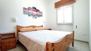 Ein Schlafzimmer mit einem hölzernen Bett, einem Nachttisch, einer Kommode, einem Fenster und einem an der Wand befestigten Kunstwerk.