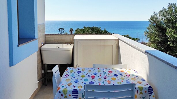 Un balcón con mesa y sillas, vista al mar y una ventana con detalles azules.
