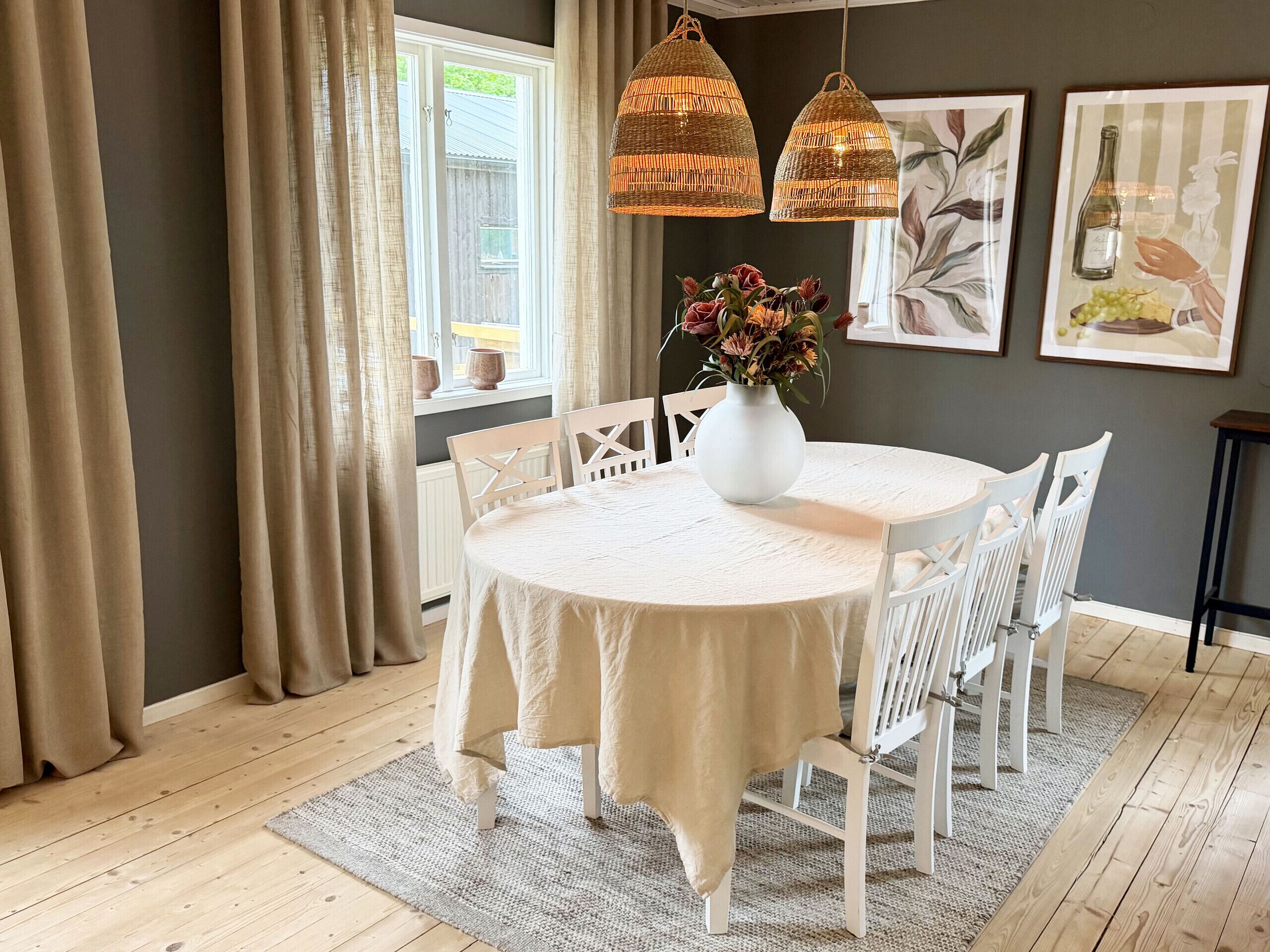 A dining area with a round table, white chairs, a vase with flowers, and hanging wicker lamps.