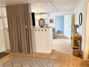 A room with a wooden floor, a beige rug, a white cabinet with hooks, a hanging light fixture, and a door leading to another room. - Rural charm and modern comfort – close to nature and swimming |SE08016 (Ljung)