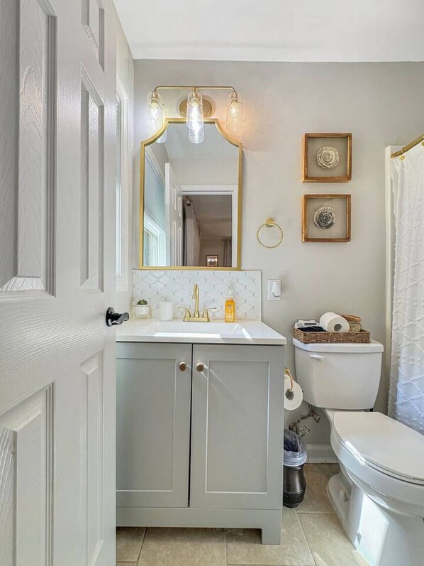 A bathroom with a white sink, a gold-framed mirror, a toilet, and framed wall art. - The Lazy Tide Inn House (Tuscaloosa)