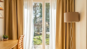 A room with a bed, a table, a chair, a lamp, and a view of a garden through a glass door.