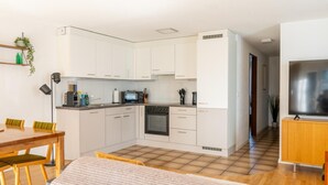 A compact kitchen with white cabinets, a microwave, stove, and a built-in oven. There is a dining area with a wooden table and chairs, and a TV on a wooden stand.