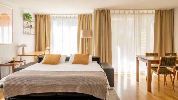 A hotel room with a bed, a desk, a chair, a lamp, and a window with curtains.