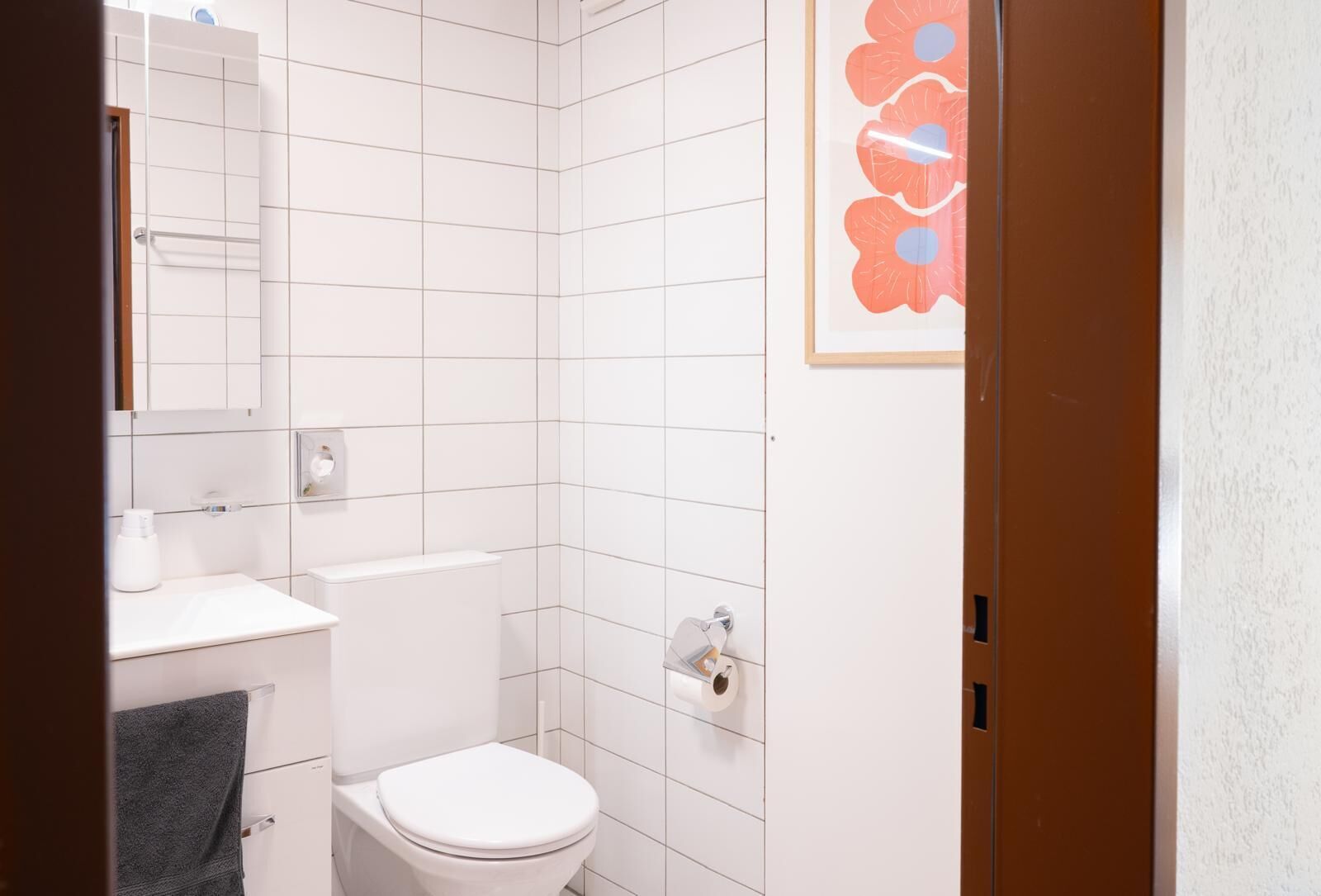 Kylpyhuoneessa on valkoiset laatat, wc-istuin, pesuallas, pyyhekoukku ja seinällä oleva kehystetty kukka-aiheinen taideteos.