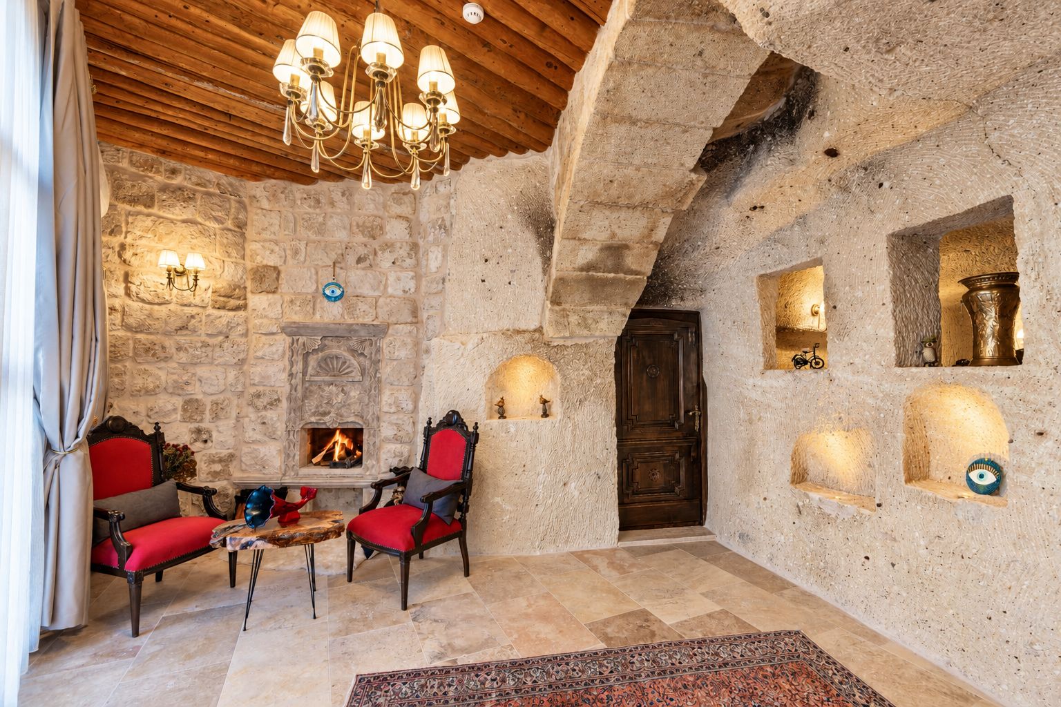 A stone-walled room with a fireplace, two red upholstered chairs, a chandelier, and a wooden ceiling.