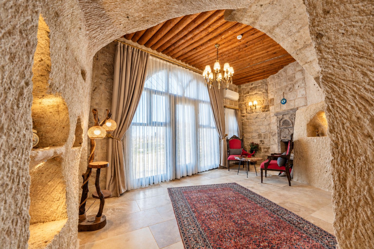 A room with stone walls, a wooden ceiling, a chandelier, a fireplace, and a red sofa.