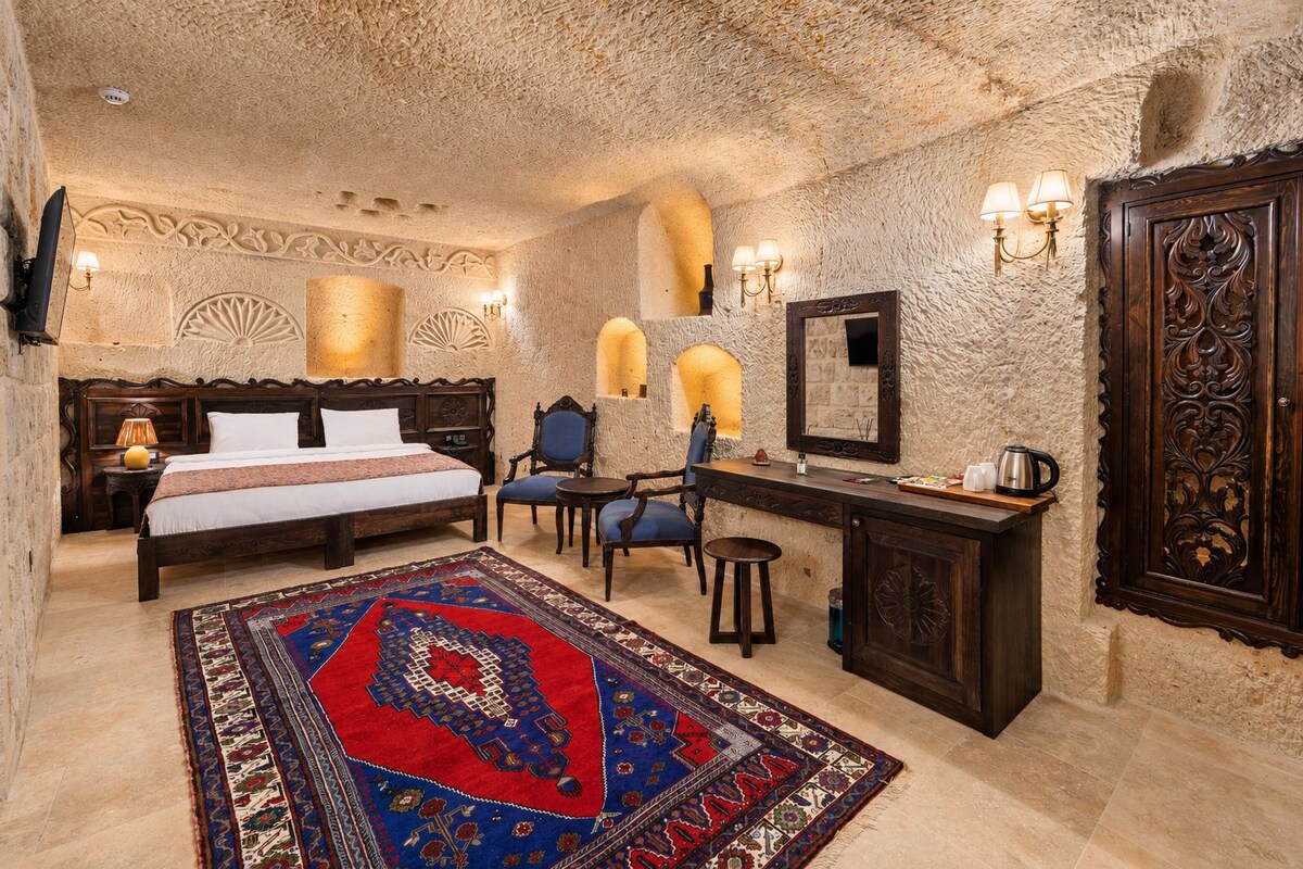 A room with a large bed, a desk, two chairs, a rug, and a wall-mounted mirror.