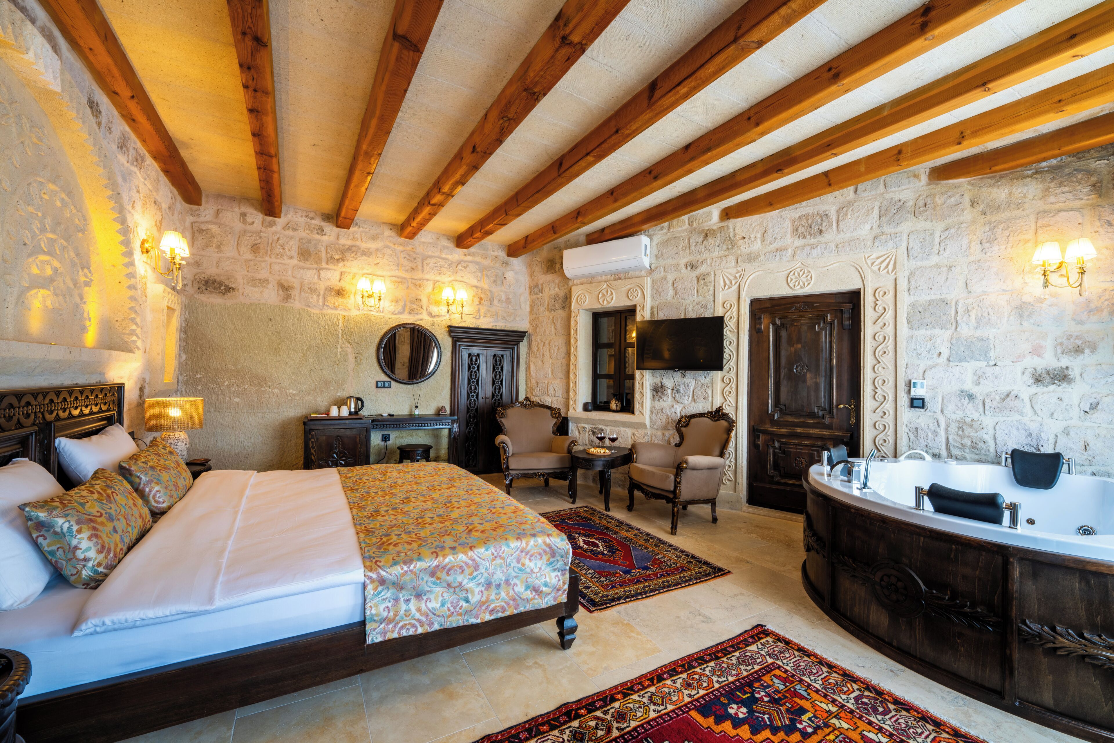 A bedroom with a bed, a sitting area with chairs, a TV, a large bathtub, and stone walls.