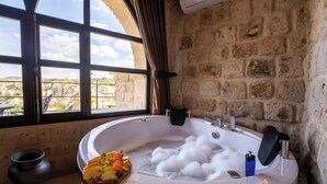 A room with a stone wall, a large window, a bathtub with bubbles, a wooden tray with fruits, and a candle. - Aurelia Cave Cappadocia (Urgup)