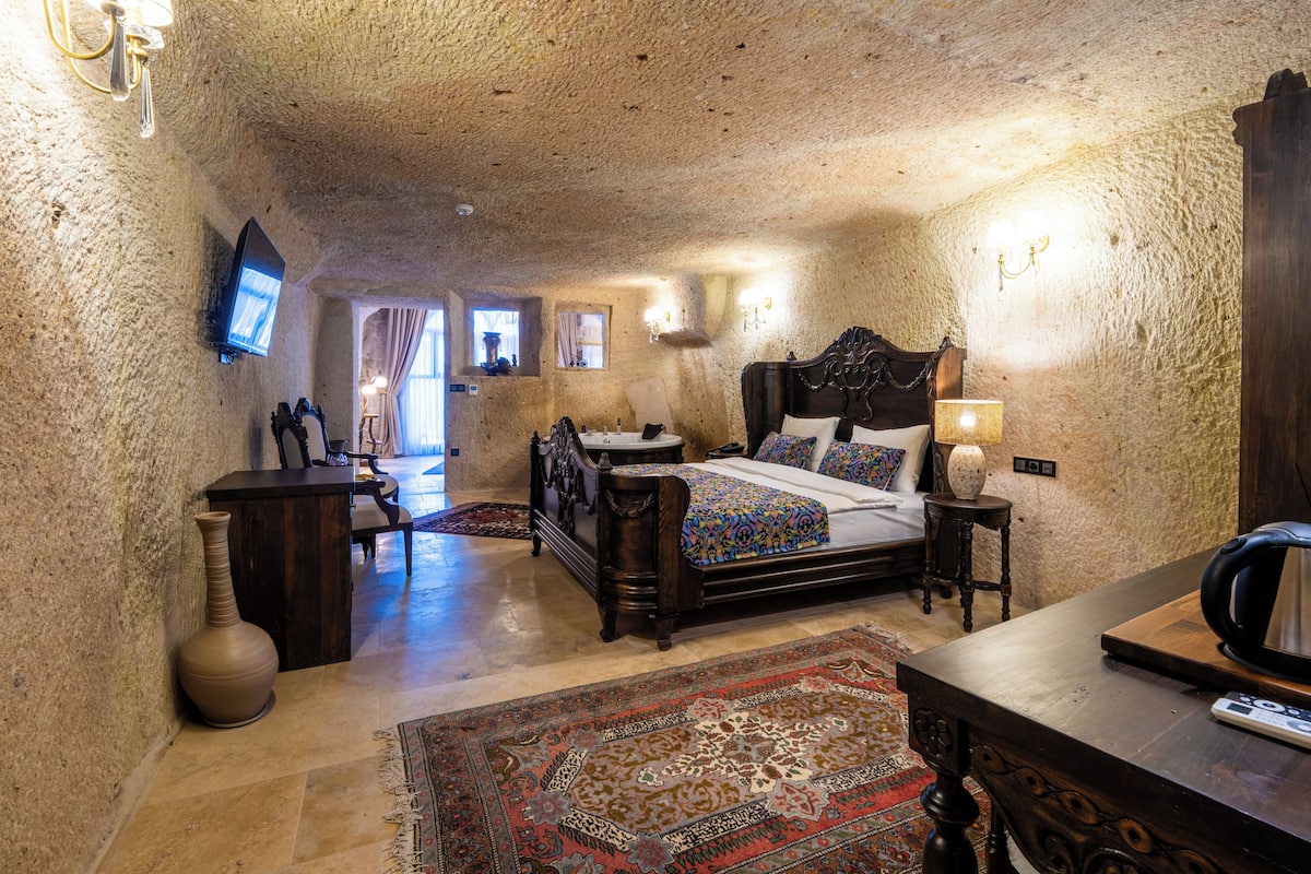 Quarto com parede de pedra, cama, escrivaninha, cadeira, televisão e luminária.