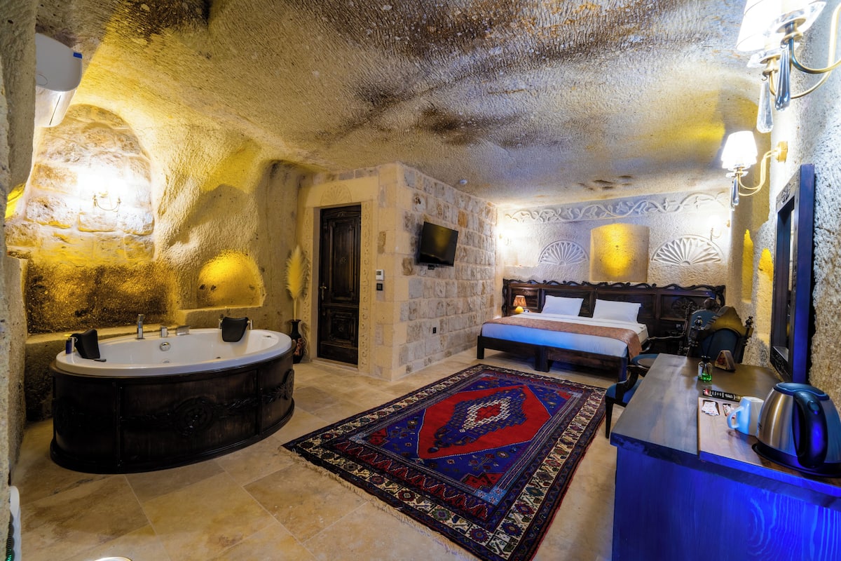Quarto com parede de pedra, banheira, cama, televisão e escrivaninha com chaleira.