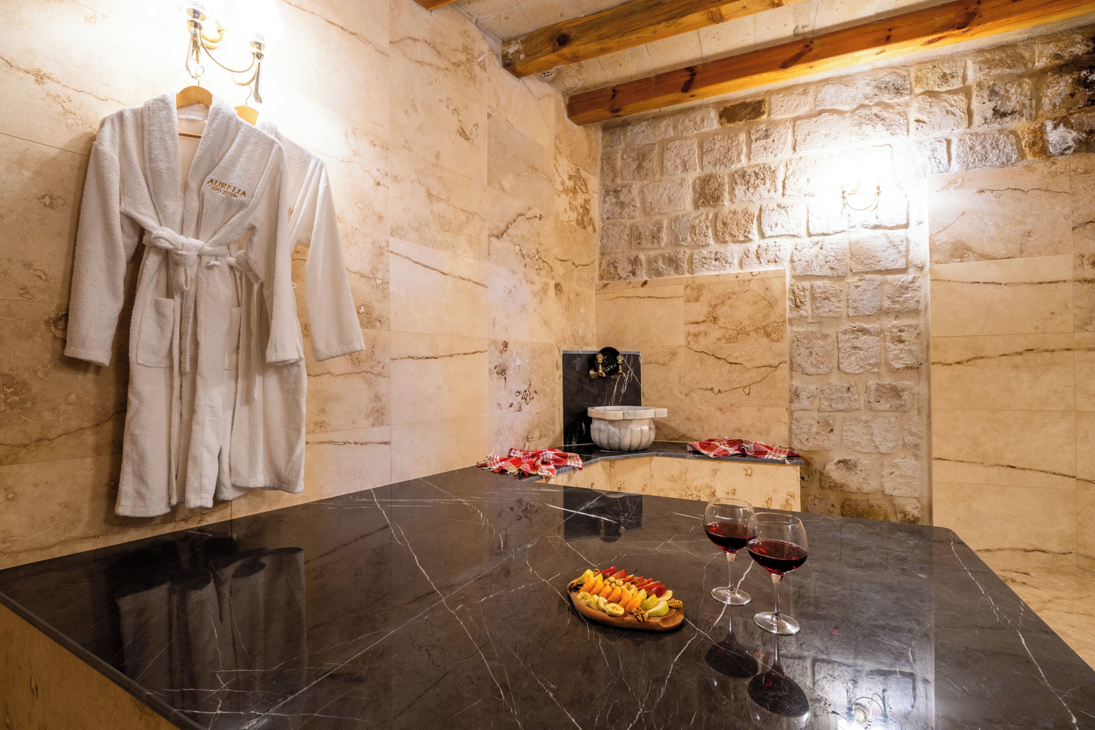 A marble-topped table with a fruit platter and wine glasses, a robe hanging on the wall, and a stone wall with a sink.