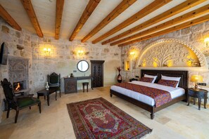 Un dormitorio con pared de piedra, vigas de madera, chimenea, cama con ropa de cama estampada y mesita de noche con lámpara.