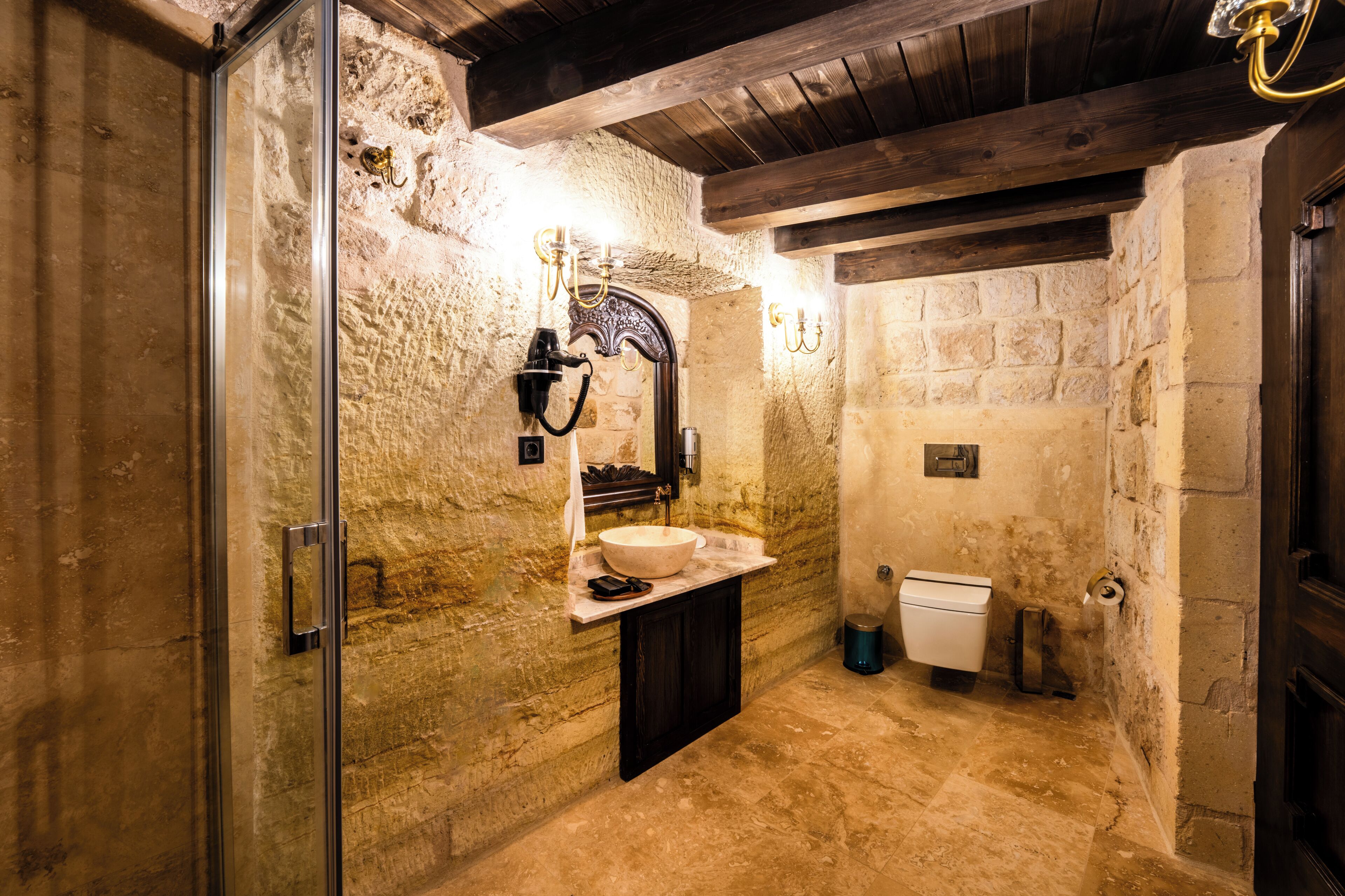 A stone-walled bathroom with a wooden ceiling, a sink, a toilet, and a shower area.