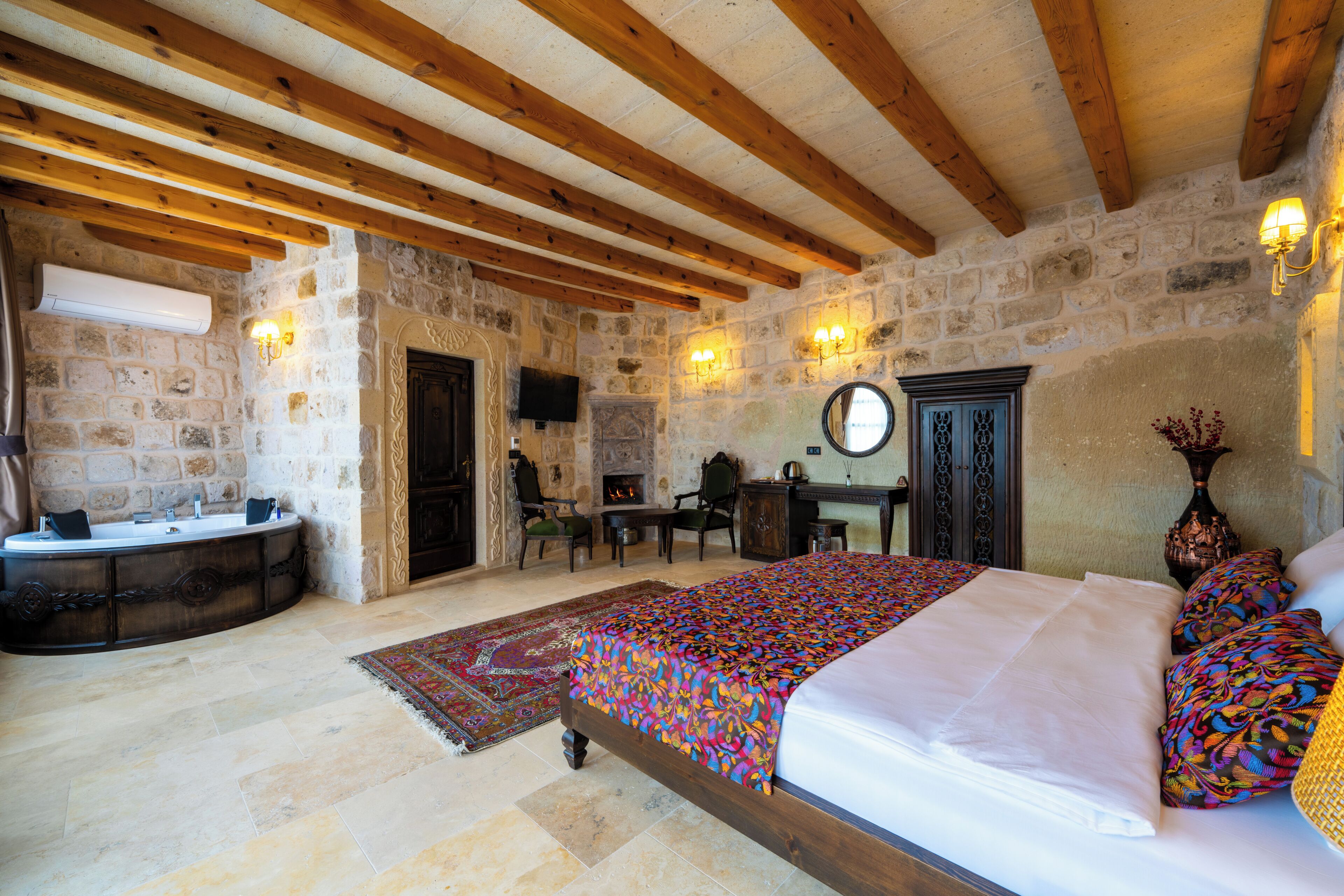 A bedroom with a stone wall, wooden beams, a bed with colorful bedding, a desk, and a fireplace.