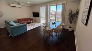 A living room with a sofa, a glass table with chairs, a flat-screen TV, and a large window with a view of buildings. - Private Balcony | Gorgeous Center City Bilevel Apt (Philadelphia)