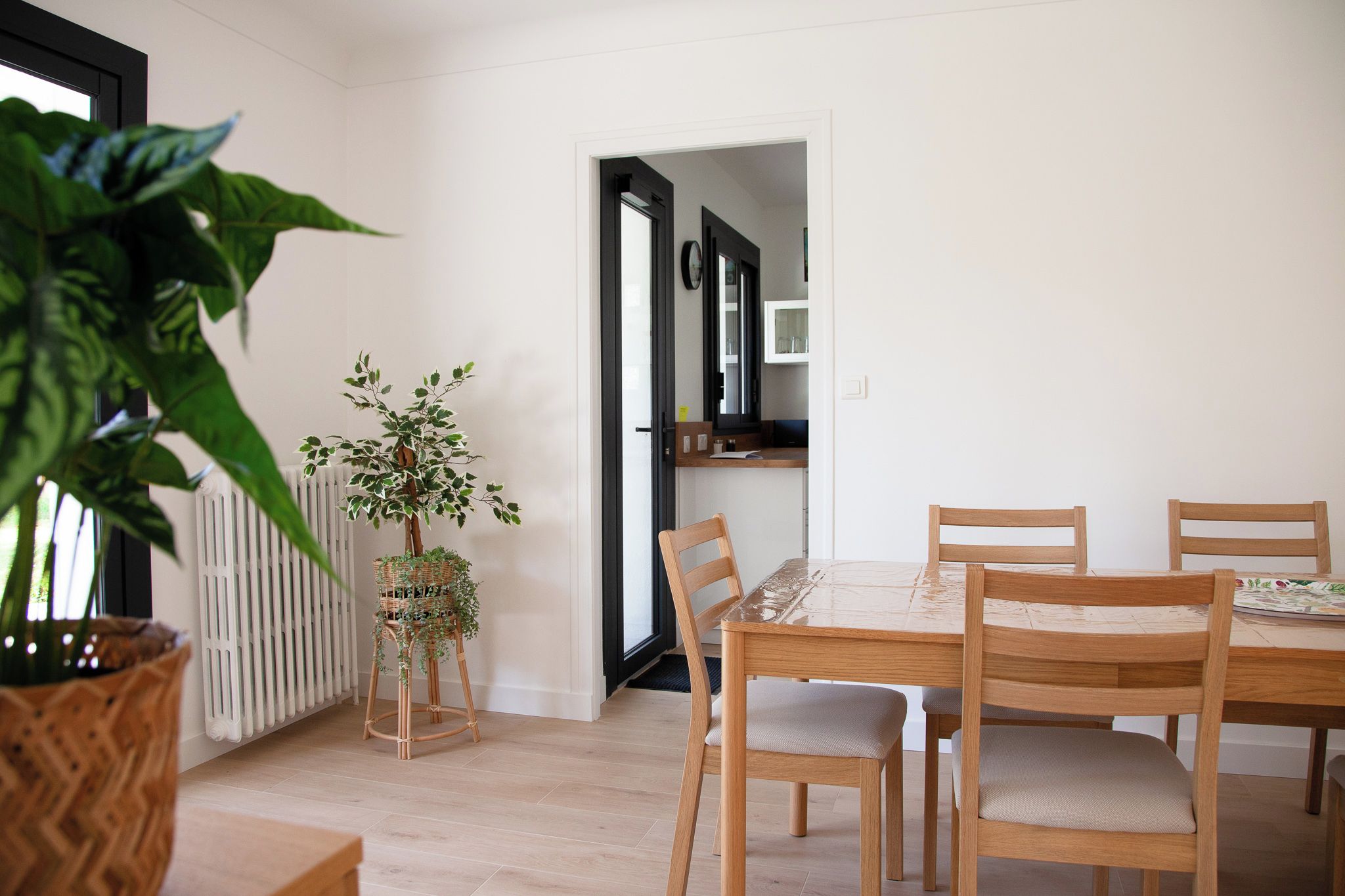 Un coin repas avec une table et des chaises, un radiateur, une plante en pot et une porte-fenêtre donnant sur une autre pièce.