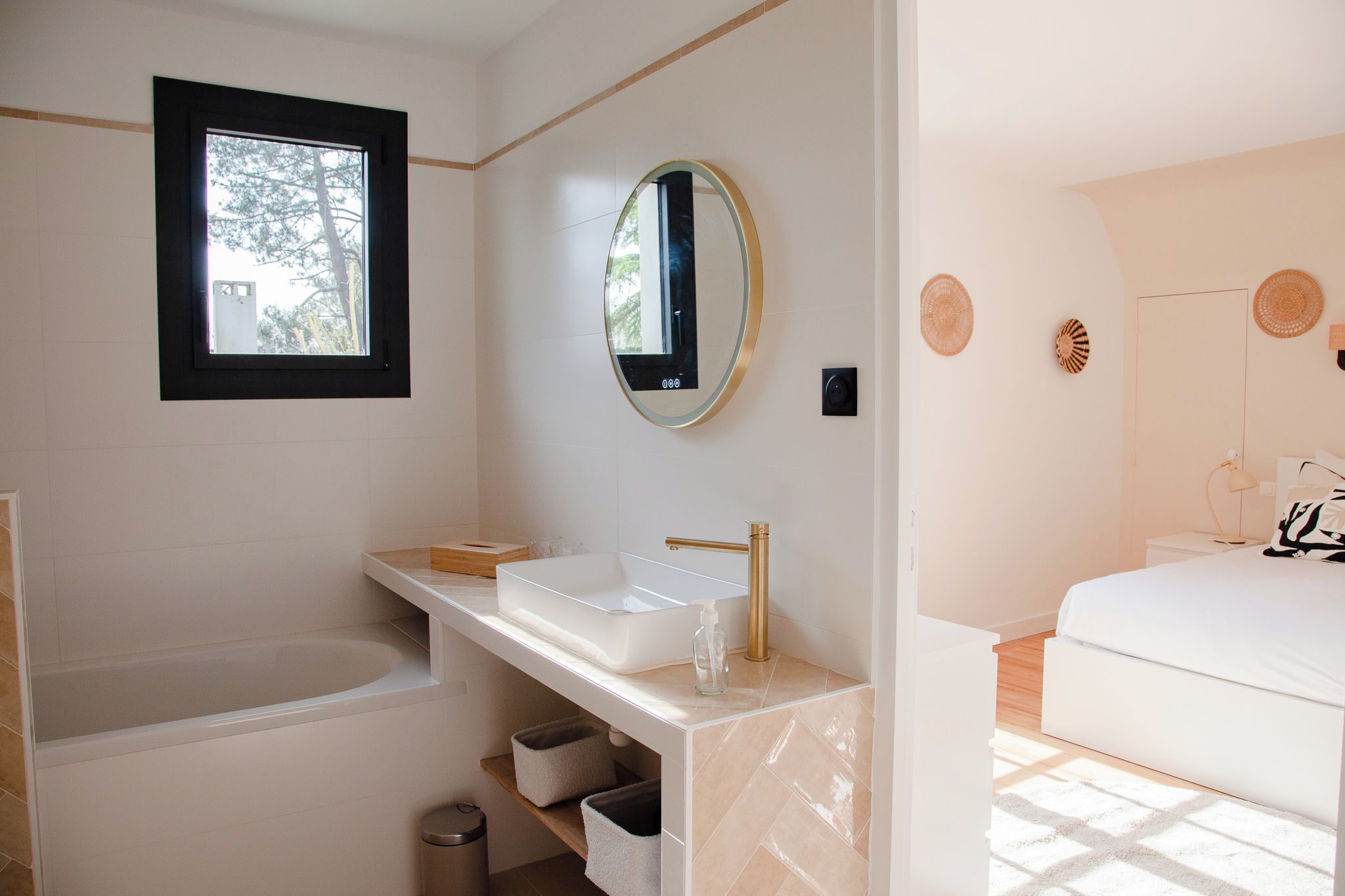 A modern bathroom with a bathtub, a sink with a gold faucet, and a large mirror. There is a window above the sink and a door leading to another room.