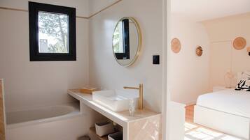 A modern bathroom with a bathtub, a sink with a gold faucet, and a large mirror. There is a window above the sink and a door leading to another room.