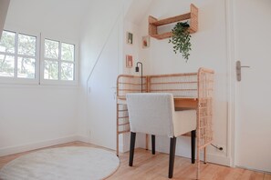 Ett rum med en vit vägg, ett fönster, en vit dörr, en flätad stol, ett litet bord och en hängande växt.