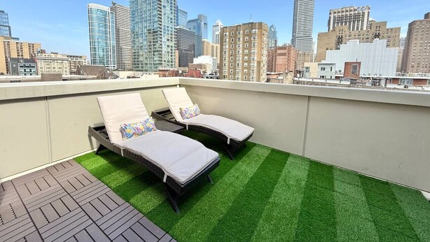Une terrasse sur le toit, dotée de deux chaises longues et d’un gazon synthétique, offrant une vue sur un paysage urbain composé de différents bâtiments.