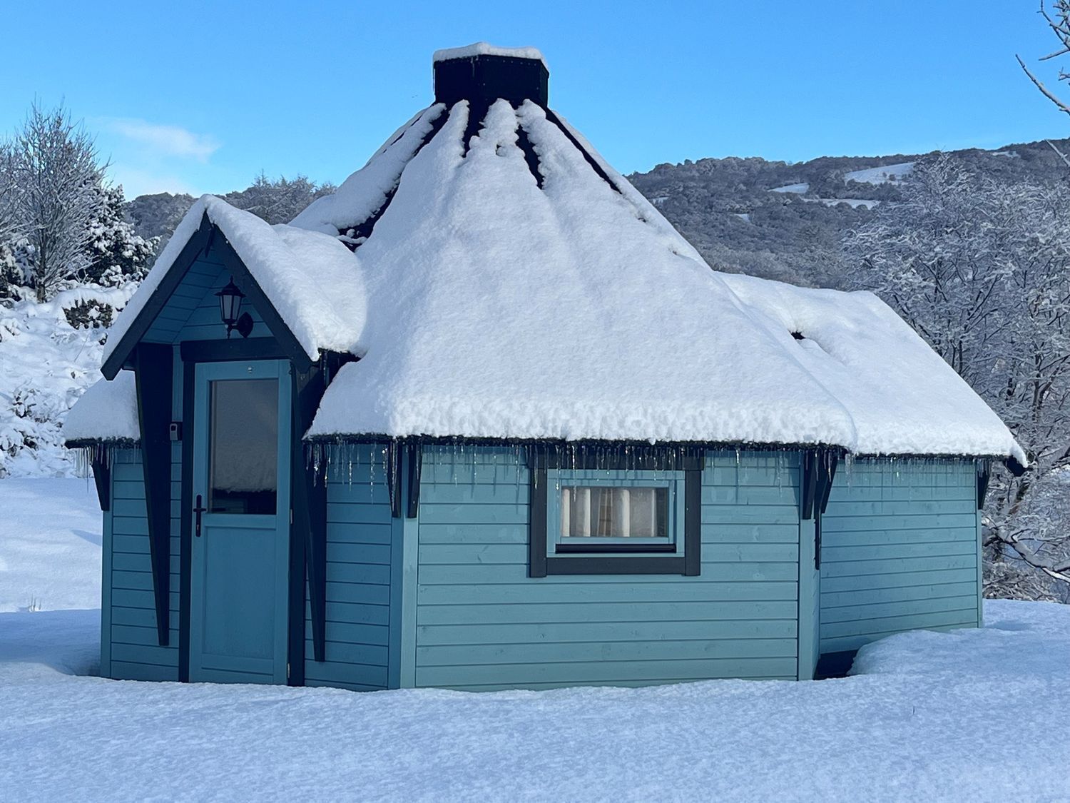 A small, single-story house with a steeply pitched roof covered in snow, a small window, and a door.