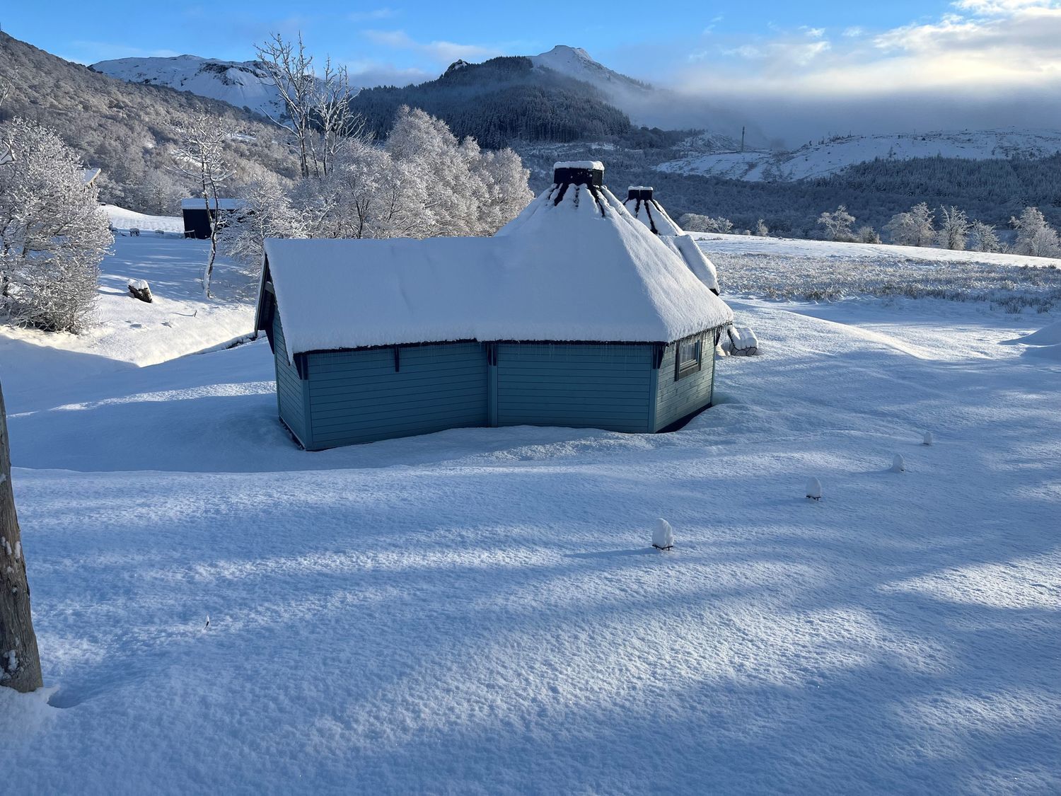 Una cabaña cubierta de nieve con un techo inclinado, rodeada de un paisaje nevado y montañas.