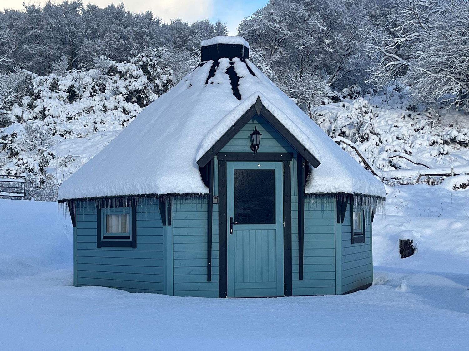 A small, light blue cabin with a snow-covered roof and a single door, surrounded by a snowy landscape.