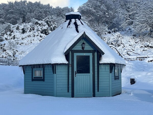 A small, light blue cabin with a snow-covered roof and a single door, surrounded by a snowy landscape. - Little Luxe Lodges 1 (Kyle)
