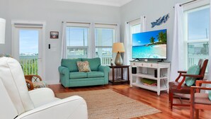 A living room with a teal sofa, white armchair, wooden floor, TV, and a view through the window. - Rhapsody in Blues (Port St. Joe)
