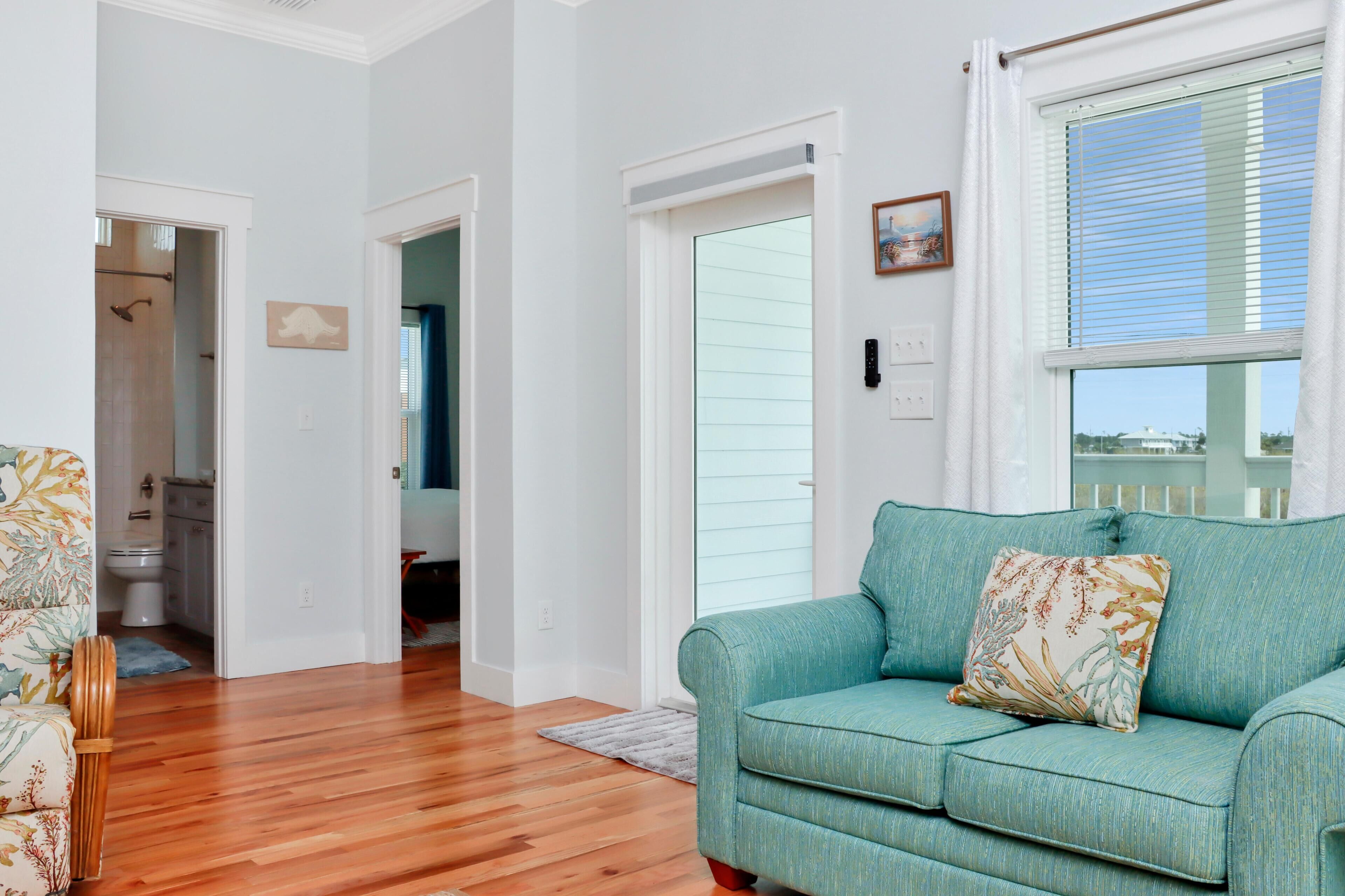 A living room with a teal sofa, wooden flooring, a door leading to a bathroom, and a window with blinds.