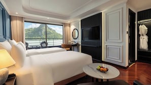 Ein Hotelzimmer mit Bett, Schreibtisch, Fernseher, rundem Tisch mit Obst und Blick auf Berg und Wasser.