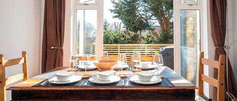 A dining table set for four with chairs, a view of trees and a fence through large windows, and a wooden floor.