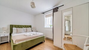 Ein Schlafzimmer mit einem grünen, bezogenen Bett, weißen Nachttischen, Holzdielen, einem Fenster mit Vorhängen, einem weißen Kleiderschrank und einem Spiegel.