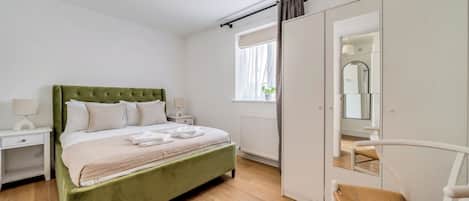 A bedroom with a green upholstered bed, white nightstands, a wooden floor, a window with curtains, a white wardrobe, and a mirror.